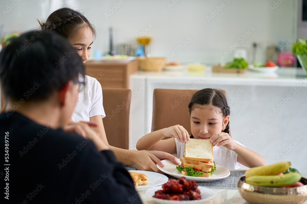 Daughter is eating sandwich on home dinner table with parent