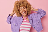 Optimistic curly woman keeps hands on head smiles broadly keeps eyes closed has fun dressed in casual striped t shirt and purple jacket expresses positive emotions isolatated over pink background.