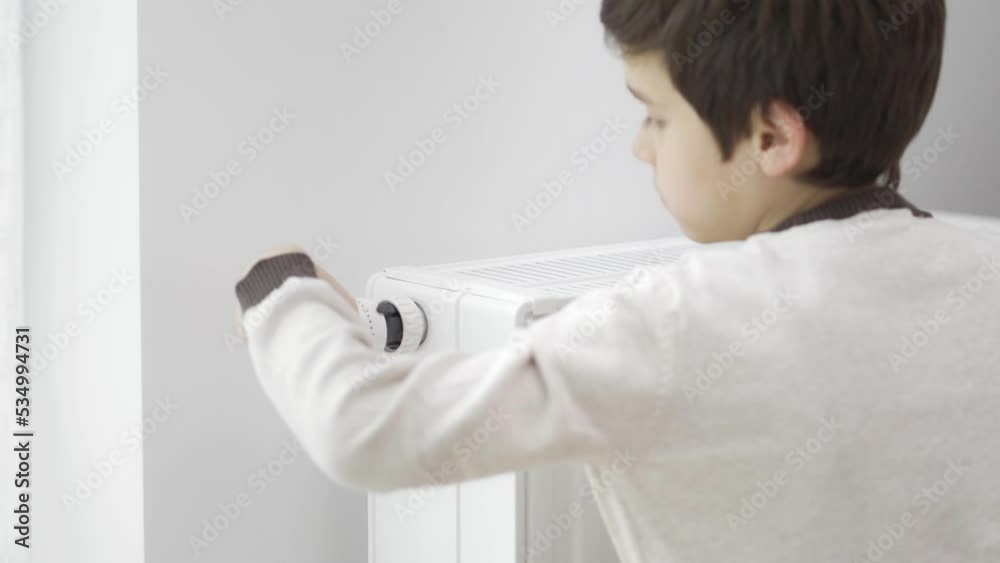 The child touches the cold radiator of the home heater with frozen hands and increases its heating power by the maximum value to warm up. The boy hugs the heater. Heating during the energy crisis. The child touches the cold radiator of the home heater with frozen hands and increases its heating power by the maximum value to warm up. The boy hugs the heater. Heating during the energy crisis.