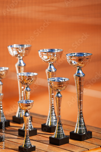 A lot of silver plated championship cups prepared for the winners of a tennis sport competition against clay tennis court in background. Symbol for victory.
