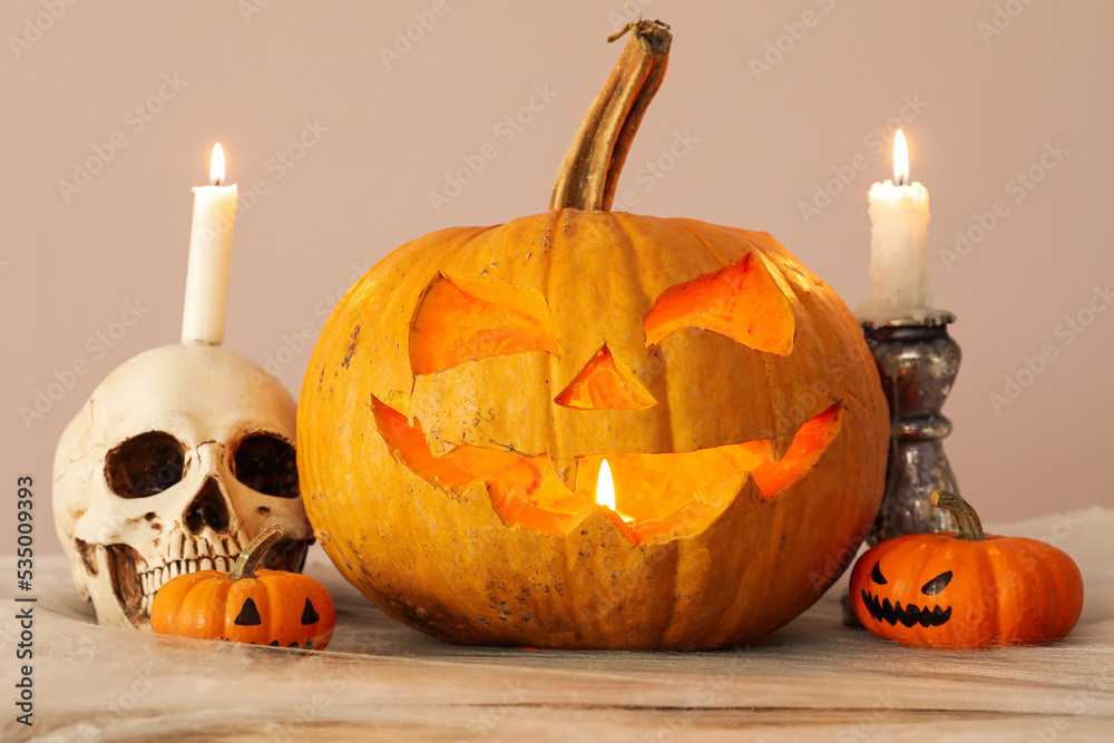 Jack-O-Lantern pumpkin with burning candles and human skull on table near beige wall