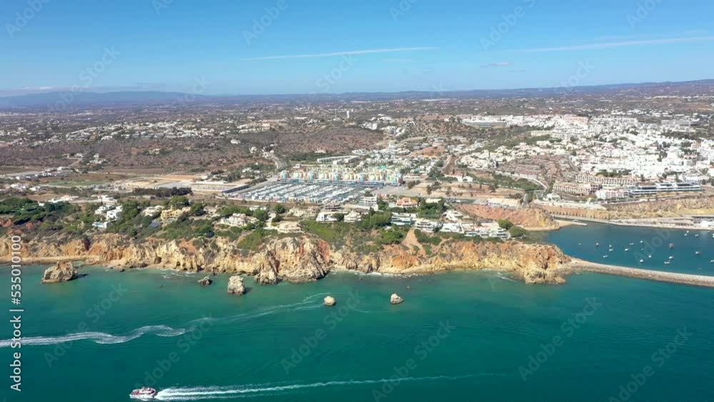 Cinematic high angle view of Albufeira landscape with high rocky shore and luxury houses on top. Turquoise water colour. Tourist boats passing. In background the harbour of Albufeira. Drone forward Cinematic high angle view of Albufeira landscape with high rocky shore and luxury houses on top. Turquoise water colour. Tourist boats passing. In background the harbour of Albufeira. Drone forward