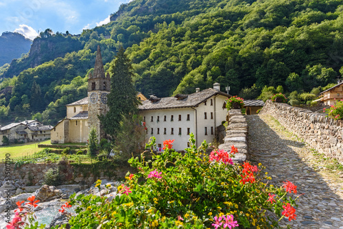 Lillianes, Aosta Valley. Italy. View of the stone bridge over the Lys stream and the Church of San Rocco. Some flowers in foreground. July 27, 2022.