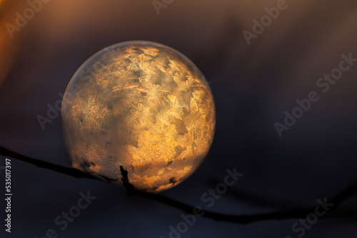 Close-up of frozen bubble against sun during sunset in orange light on branch with small crystals