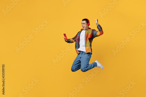 Full body young fun happy cool middle eastern man wears casual shirt white t-shirt headphones listen music jump high do winner gesture hold use mobile cell phone isolated on plain yellow background.