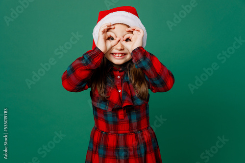 Merry cheerful funny cool happy little child kid girl 6-7 years old wearng red dress Christmas hat posing holding hands near eyes isolated on plain dark green background. Happy New Year 2023 concept.