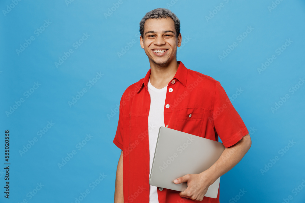 Young fun happy IT man of African American ethnicity 20s wear red shirt hold use work on laptop pc computer look camera isolated on plain pastel light blue cyan background. People lifestyle concept