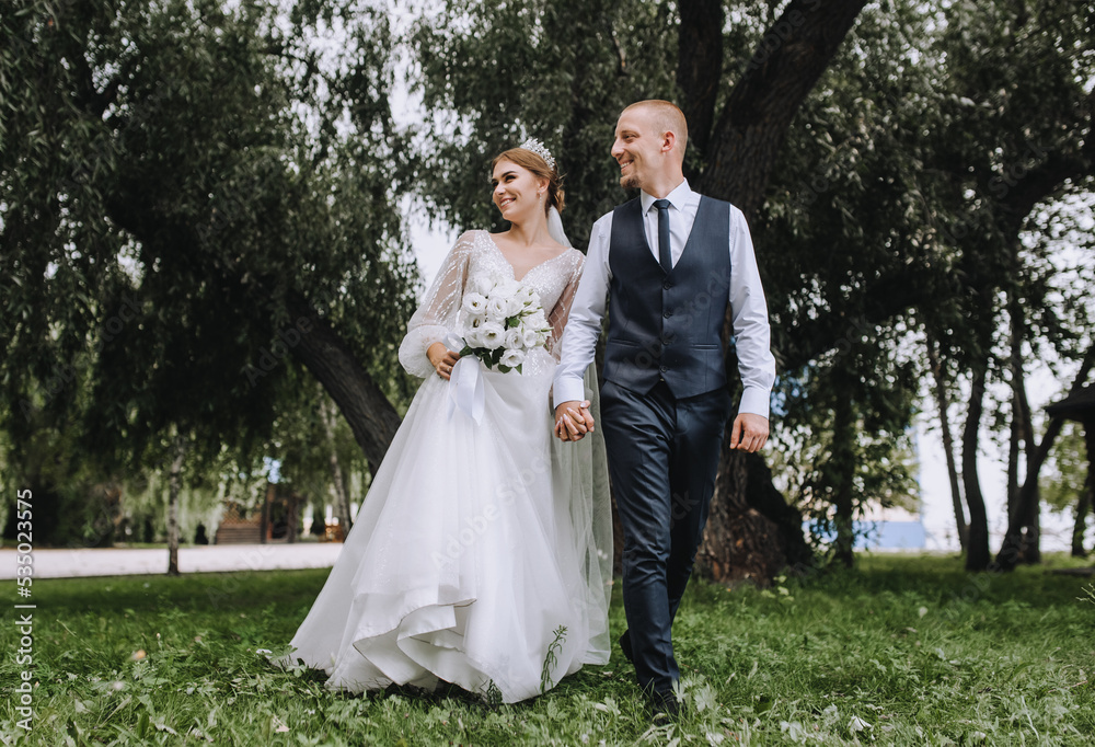 A stylish groom in a blue vest and a beautiful smiling bride are walking in a park in nature on green grass, holding hands. Wedding photo of newlyweds in love.