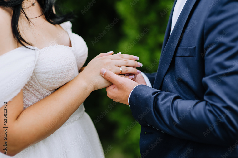 The groom in a blue suit and the bride in a white dress stand at the ceremony holding hands with a gold ring on their finger. Close-up wedding photography, portrait.