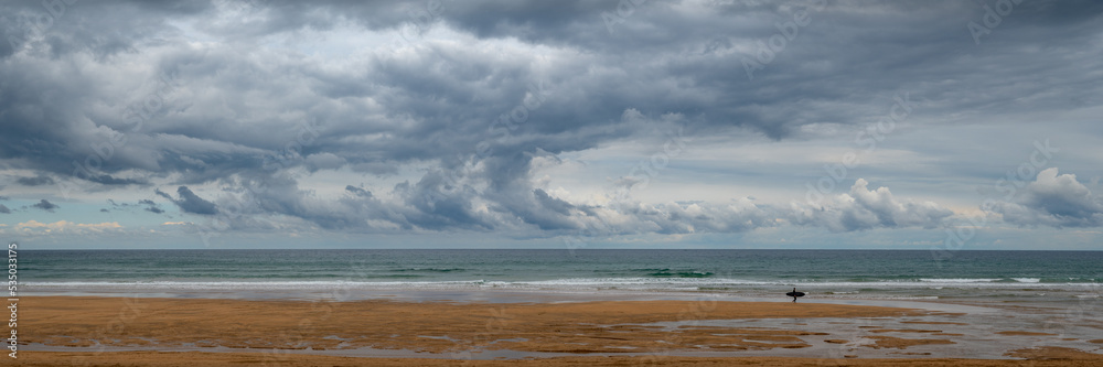 Epic Panorama of a Beach at the Atlantic Coast. The Silhouette of single Surfer with his Board against a dramatic Sky
