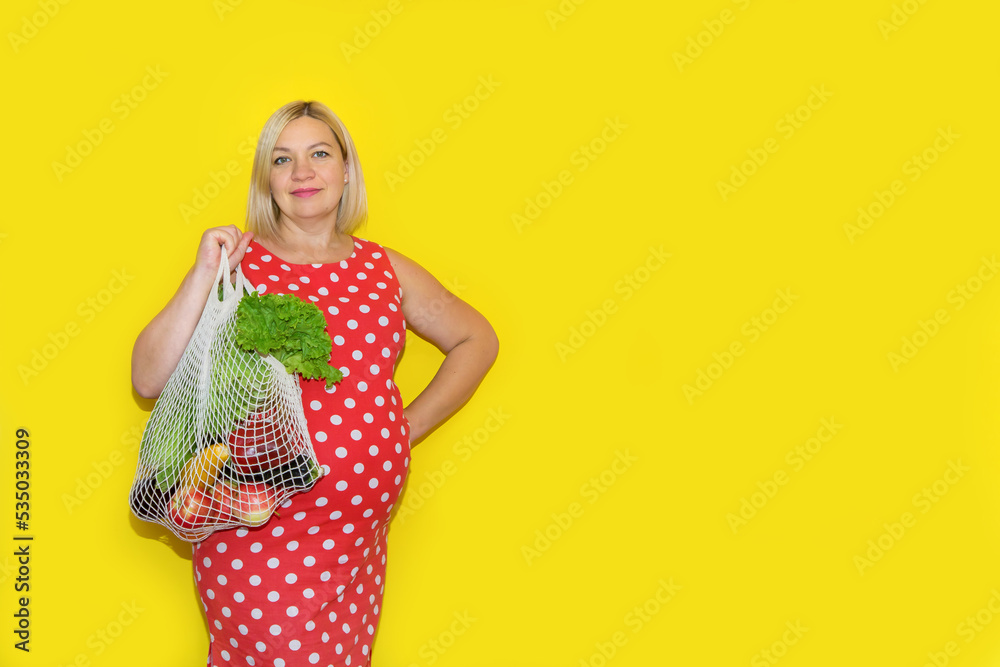 pregnant woman in a red dress with a shopping bag of vegetables and fruits on a yellow background with copy space