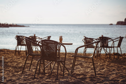 empty cafe table and chairs on sandy beach in front of sea golf with clear sky before sunset