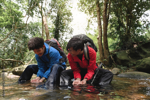 Two young trekkers or traveler sits on rocks in mountain river and enjoys with mountain stream water in hands while washing face with natural water. Refresh himself after long journey.