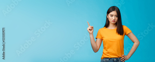 Moody upset asian girlfriend, sulking look offended and displeased pointing upper left corner, pouting disappointed, standing distressed blue background wear yellow t-shirt