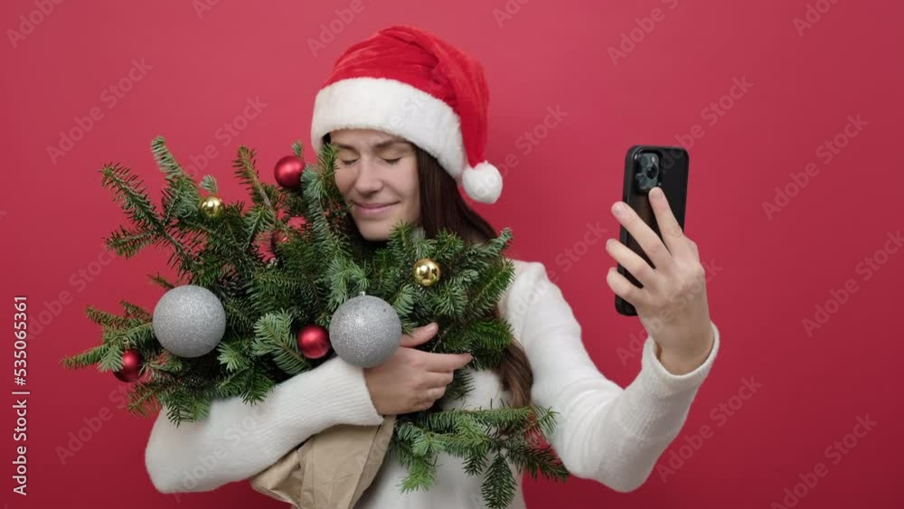 Portrait of good-looking young brunette female in Santa Claus hat holding bouquet of spruce branches do selfie shot on mobile cell phone post photo on social network, posing over red studio background Portrait of good-looking young brunette female in Santa Claus hat holding bouquet of spruce branches do selfie shot on mobile cell phone post photo on social network, posing over red studio background