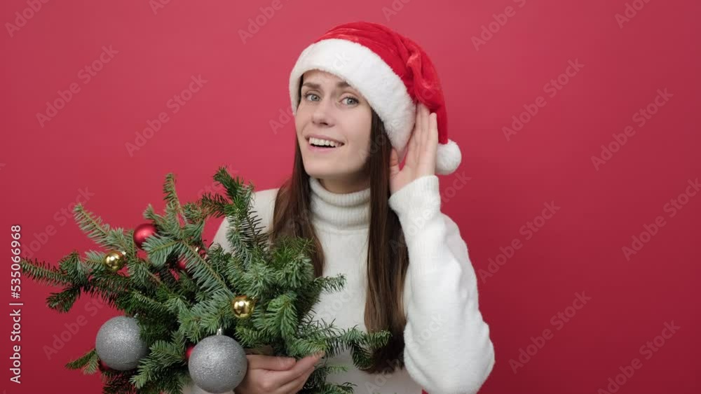 Curious nosy young woman wears white sweater and Santa hat holding bouquet of spruce branches try to hear you overhear listening intently, posing isolated over red color background wall in studio Curious nosy young woman wears white sweater and Santa hat holding bouquet of spruce branches try to hear you overhear listening intently, posing isolated over red color background wall in studio