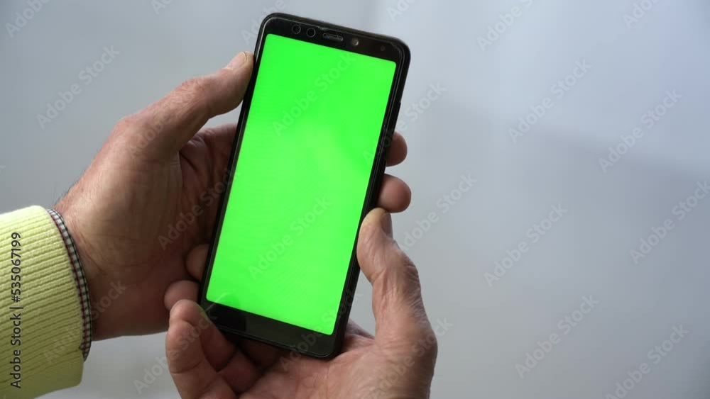 Old man holding smartphone with chromakey green screen vertically in wrinkled hands against white background at home Old man holding smartphone with chromakey green screen vertically in wrinkled hands against white background at home