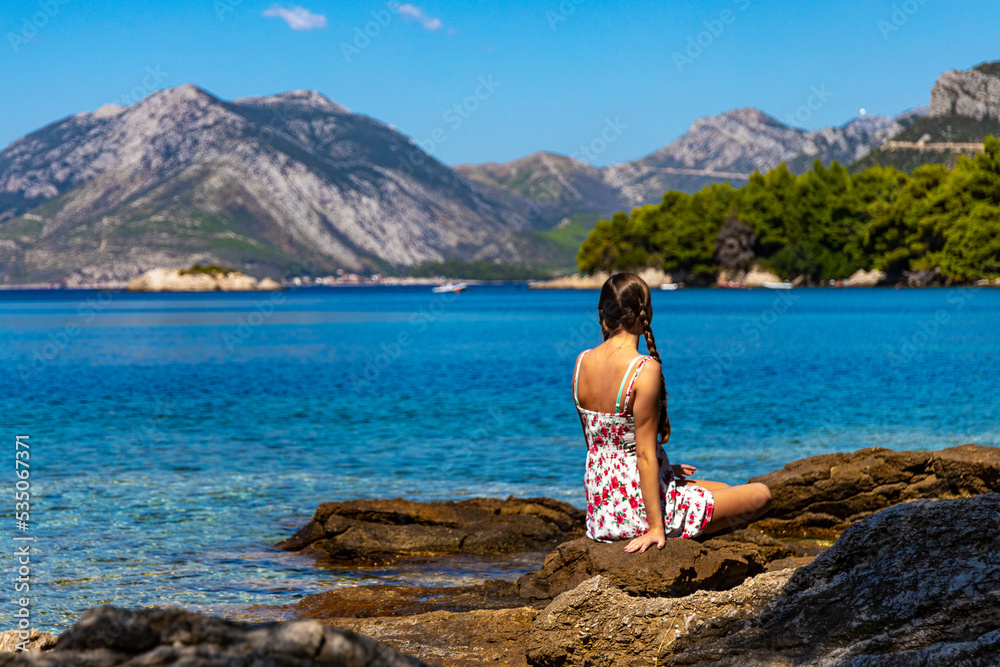 beautiful girl in a dress sits on the rocks on the seashore in croatia with a view of the mighty mountains in the background; vacation on the peljesac peninsula by the mediterranean sea
