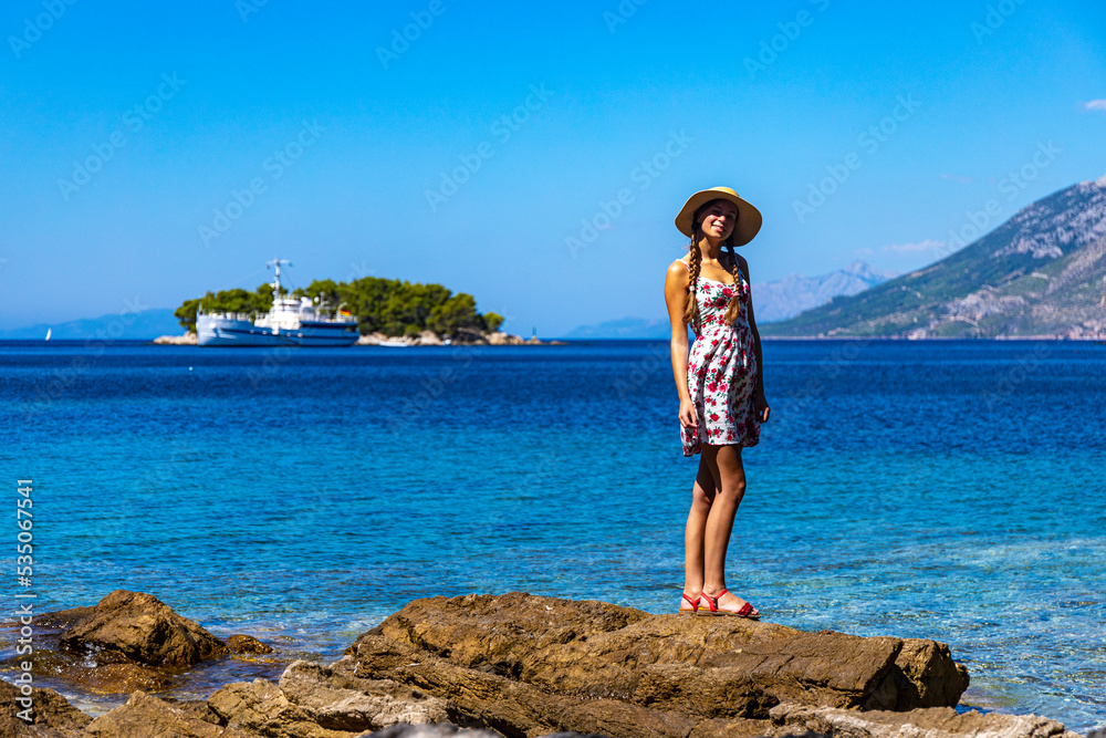 beautiful woman in a dress and hat enjoying a sunny day by the sea in croatia, paradise beach on the peljesac peninsula with islands and a ship in the background