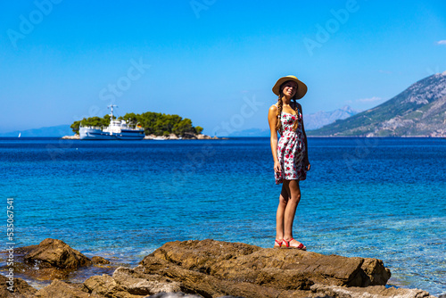 beautiful woman in a dress and hat enjoying a sunny day by the sea in croatia, paradise beach on the peljesac peninsula with islands and a ship in the background