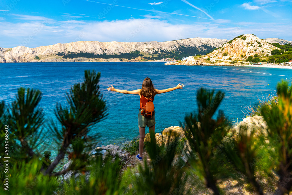 a beautiful girl stands over the rocky coast of croatia with her hands raised in the air; active vacation in croatia, hiking on the mediterranean sea, krk island