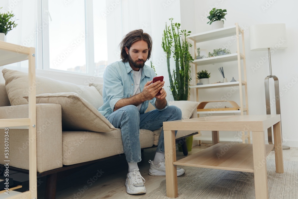 A freelance man in a white T-shirt, blue jeans, and shirt sits on the couch with his phone at home on his day off and plays games