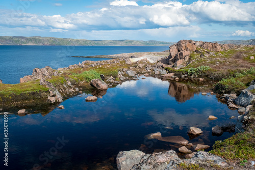 Natural pool in the rock. Blue sky with clouds reflected in clear water. Rocky coastline of Barents Sea near Teriberka. Scenery of Russian North. Kola Peninsula, Murmansk Oblast, Russia