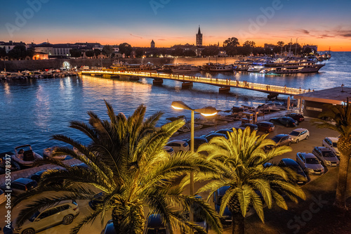 Zadar, Croatia - Aerial view of palm trees at the city of Zadar at golden sunset sky with illuminated City Bridge (Gradski most), Cathedral of St. Anastasia tower and yacht marina photo