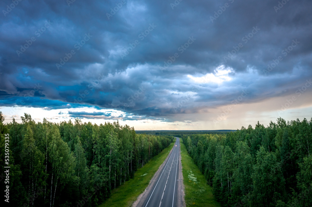 Asphalt road under the clouds. Top view of the road through the green summer forest