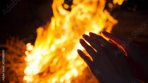 Female person tries to warm up hands near campfire flame