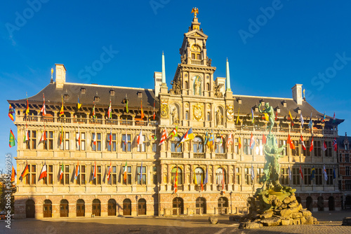 View of Brabo fountain and four-story Renaissance building of Antwerp City Hall with main facade lavishly decorated with sculptures and waving flags located on central Grote Markt square, Belgium..