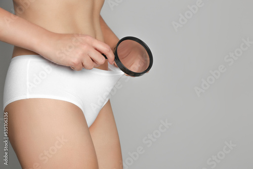 Woman examining genital herpes with magnifying glass on light grey background, closeup. Space for text