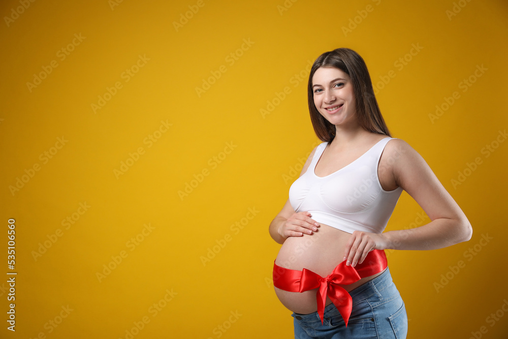 Young pregnant woman with red bow on her belly against yellow background, space for text. Time to give birth Young pregnant woman with red bow on her belly against yellow background, space for text. Time to give birth