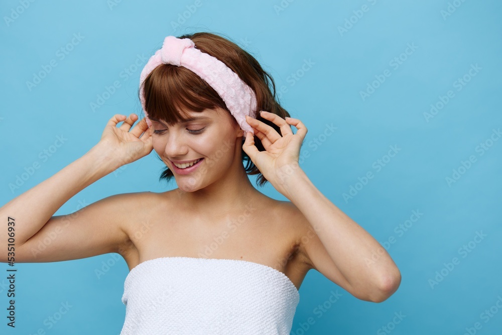 woman with smooth skin and dark beautiful hair stands on a blue background in a white towel with a pink bandage on her head after a bath with her hands near her face. Horizontal studio photography. woman with smooth skin and dark beautiful hair stands on a blue background in a white towel with a pink bandage on her head after a bath with her hands near her face. Horizontal studio photography.