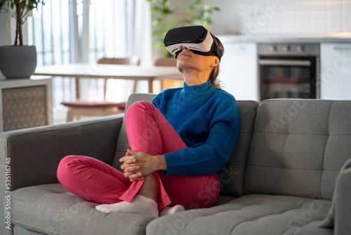 Intrigued European woman wearing virtual reality glasses relaxing after hard day at work. Girl sits on sofa next to dining room and watches video with eyes up using innovative VR and AR technologies photo