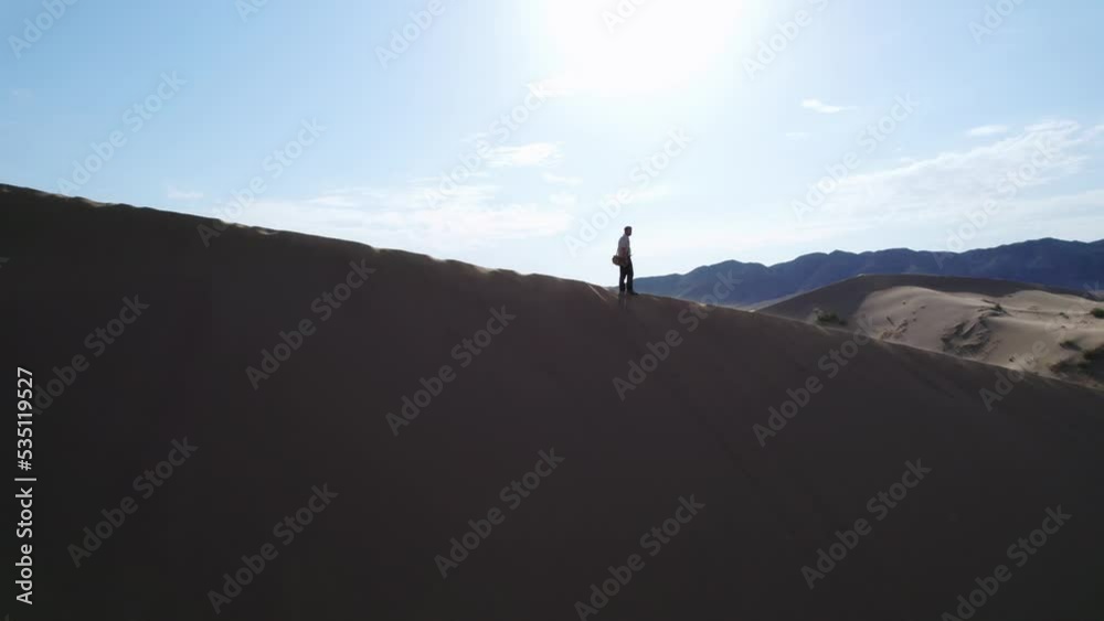 Revealing shot of the silhouette of a man on top of sand dune against blue sky and sun with behind infinite wilderness of a desert environment; aerial drone shot Revealing shot of the silhouette of a man on top of sand dune against blue sky and sun with behind infinite wilderness of a desert environment; aerial drone shot