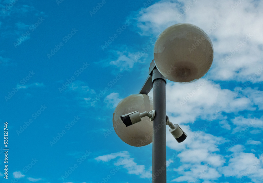 An iron pole on a city street with beautiful round lanterns and surveillance video cameras against a blue sky with white clouds. The concept of safety on the city streets. An iron pole on a city street with beautiful round lanterns and surveillance video cameras against a blue sky with white clouds. The concept of safety on the city streets.