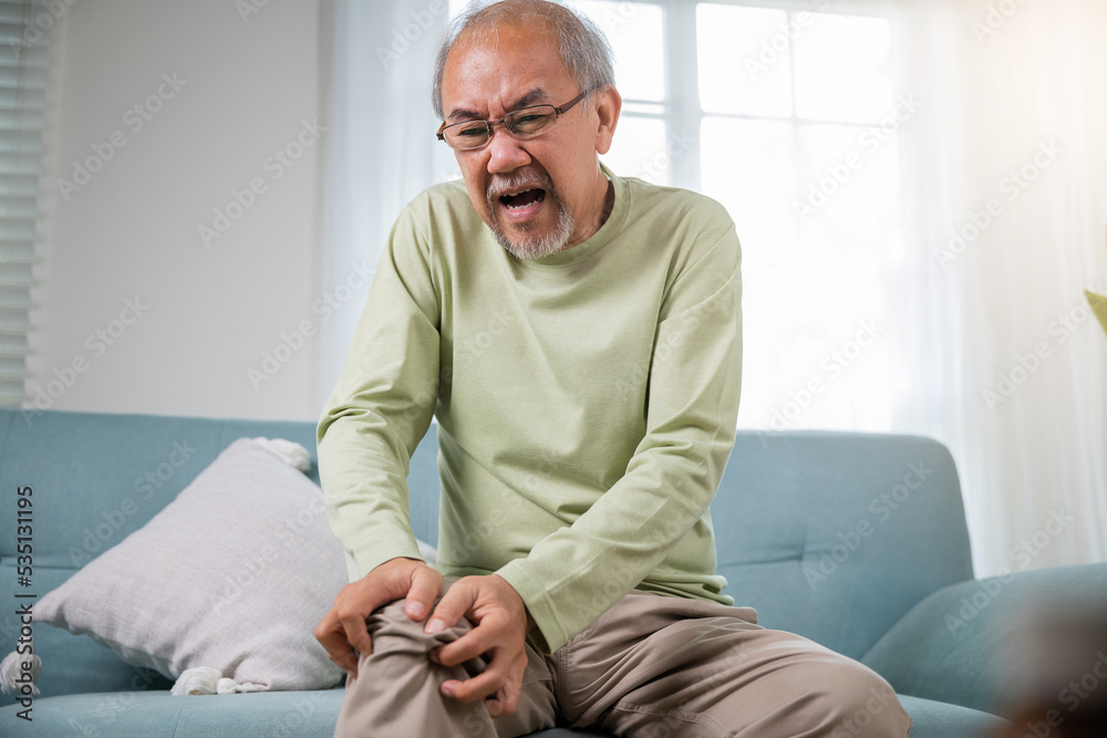 Old man suffering from knee joint pain on sofa living room, bone pain in elderly at home, senior man knee problem painful, unhappy old age hand holding on knee pain after tendon surgery
