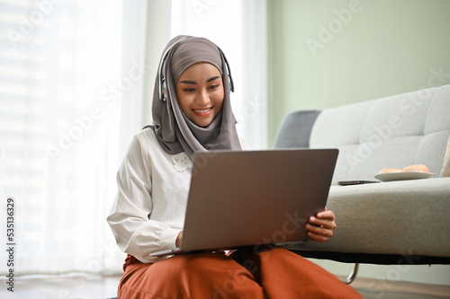 Attractive Asian Muslim woman wearing hijab, using notebook laptop in her living room