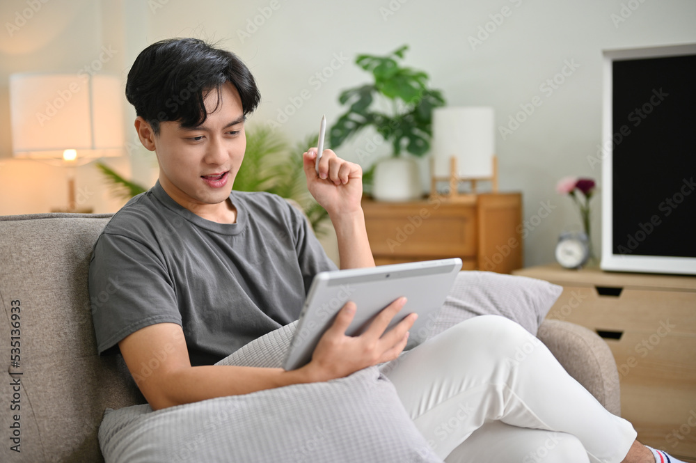 Handsome young Asian man using portable digital tablet while relaxing in his living room. Handsome young Asian man using portable digital tablet while relaxing in his living room.