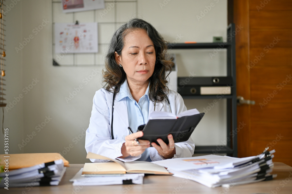 Professional senior Asian female doctor at her office desk, checking her surgery schedule. Professional senior Asian female doctor at her office desk, checking her surgery schedule.