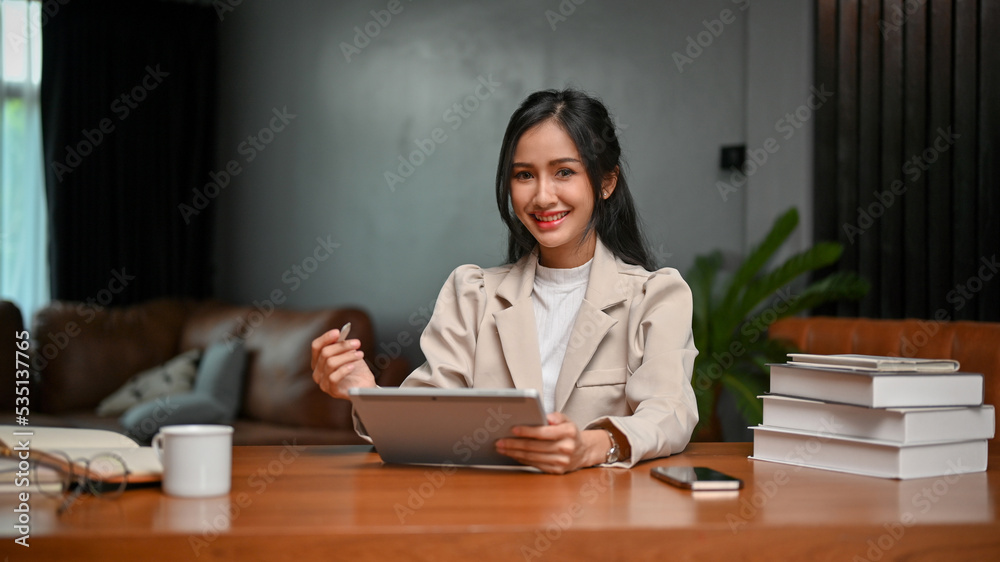 Charming Asian businesswoman using tablet at her desk, smiling and looking at the camera. Charming Asian businesswoman using tablet at her desk, smiling and looking at the camera.