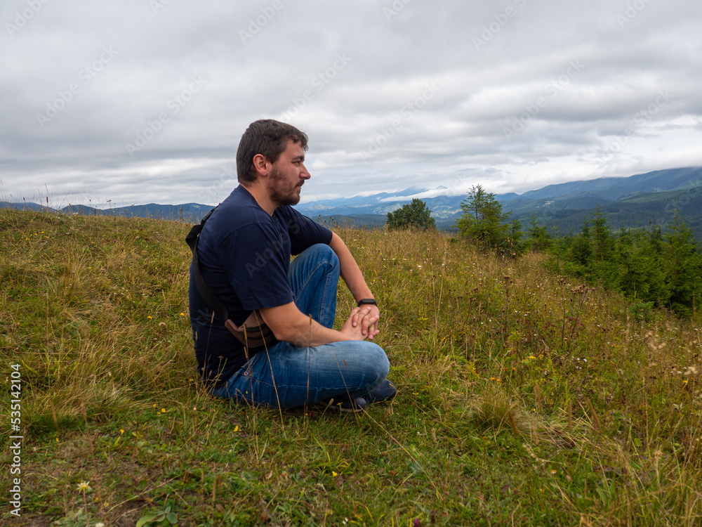 Young man admiring breathtaking view while sitting on the ground in the mountains. Man in casual clothes sits on the hill on cloudy summer day. Vacation trip to the Carpathians.