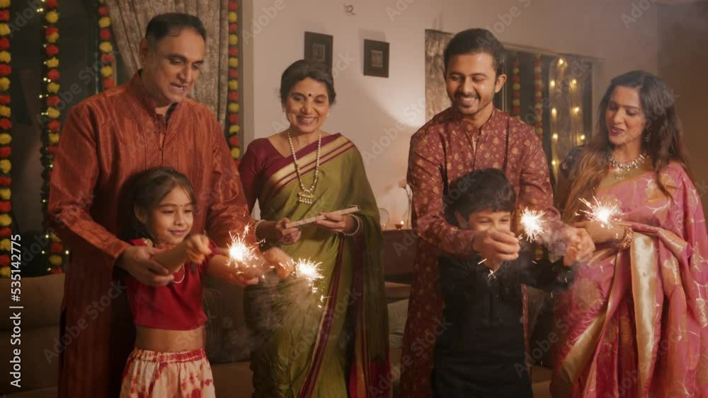 Ethnic Indian Hindu happy smiling cute young boy and a girl or kids in traditional attire playing with firecrackers or sparklers with the help of family elders or parents during Diwali festival.