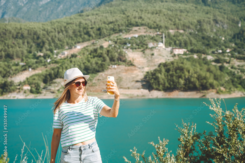 Woman taking selfie on mobile phone of mountains lake background. Traveler female having video chat on cellphone on the blue lake outdoors travel adventure vacation.
