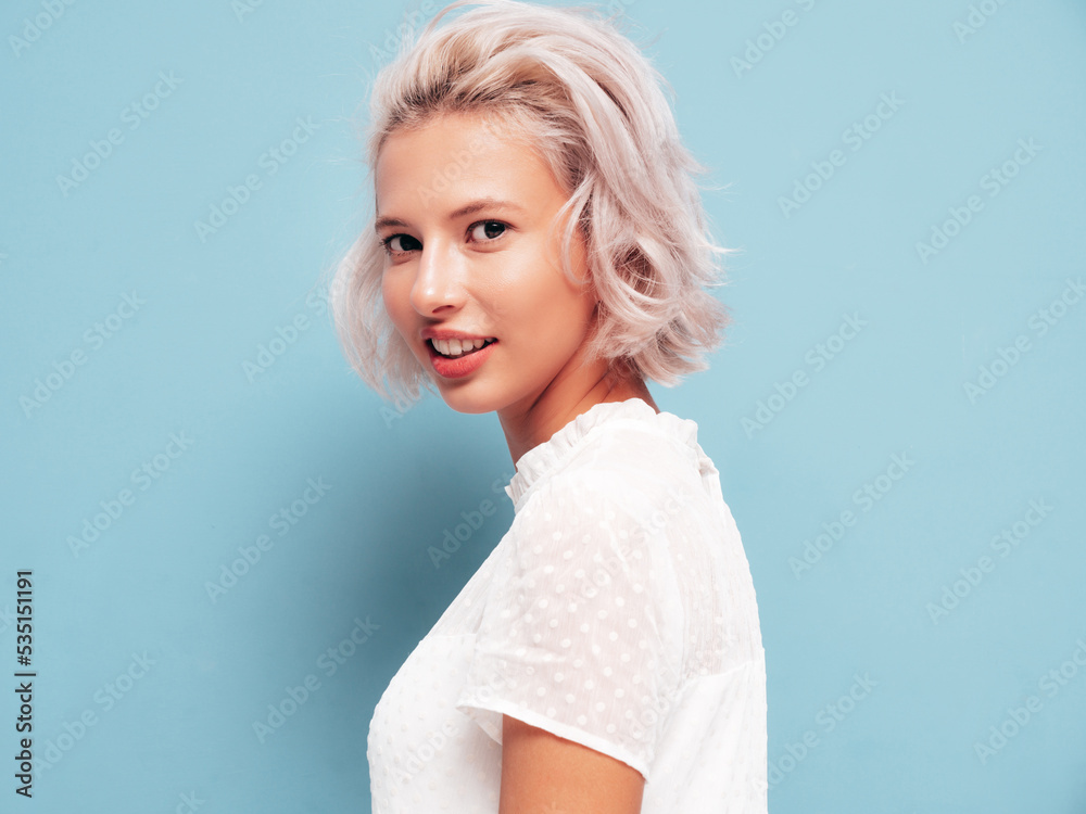 Portrait of young beautiful smiling female in trendy summer white dress. Sexy carefree blond woman posing near blue wall in studio. Positive model having fun indoors. Cheerful and happy