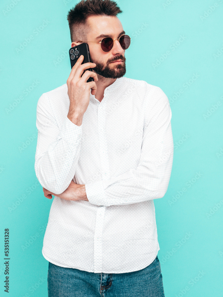 Handsome confident model.Sexy stylish man talking at smartphone. Fashion hipster male posing near blue wall in studio. Holding phone. With cellphone. In sunglasses. Isolated Handsome confident model.Sexy stylish man talking at smartphone. Fashion hipster male posing near blue wall in studio. Holding phone. With cellphone. In sunglasses. Isolated