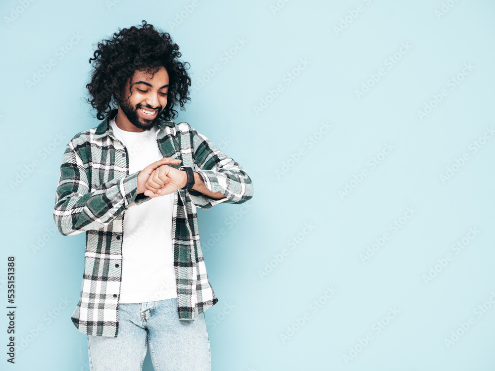 Handsome smiling hipster model.Sexy unshaven man dressed in summer stylish shirt and jeans clothes. Fashion male with long curly hairstyle posing near blue wall in studio. Looking at his watch Handsome smiling hipster model.Sexy unshaven man dressed in summer stylish shirt and jeans clothes. Fashion male with long curly hairstyle posing near blue wall in studio. Looking at his watch