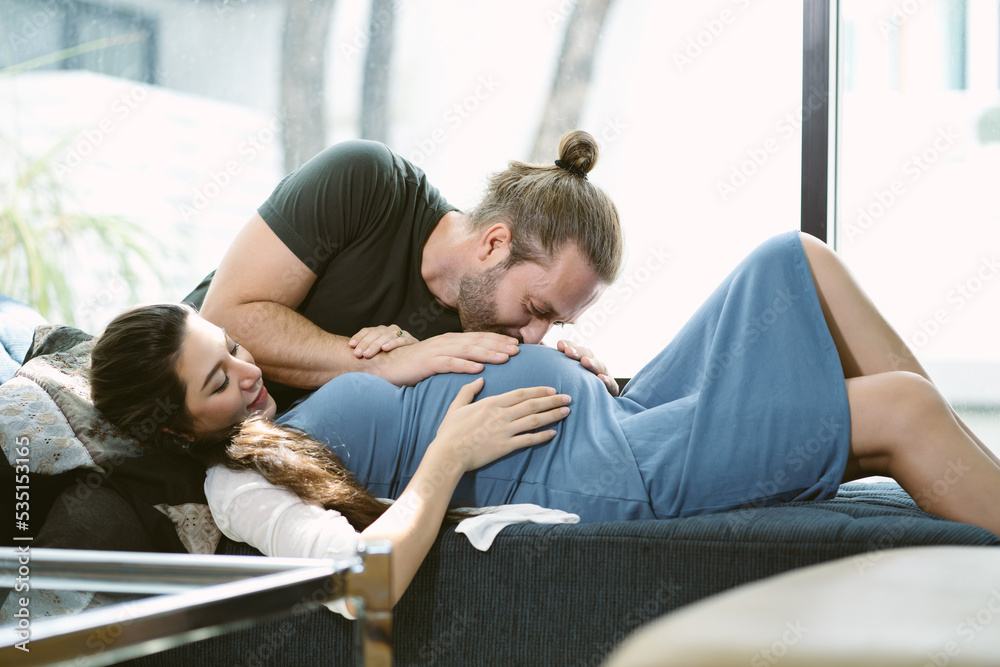 Happy handsome husband kiss pregnant wife baby in belly while his wife lying on sofa bed. Very tender moment that symbolizes the love of father to the unborn child. Maternity prenatal care concept.