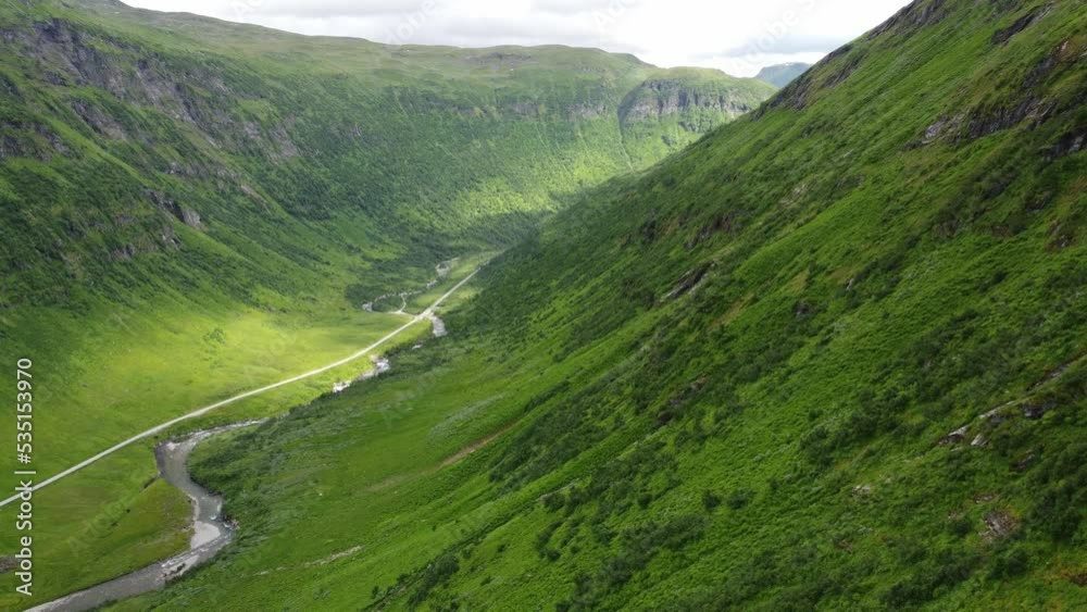 Deep lush pristine valley of Kvassdalen leading to Myrkdalen close to Vikafjell Norway - Aerial following green mountainside downwards while looking at sunrays hitting river and road in bottom Deep lush pristine valley of Kvassdalen leading to Myrkdalen close to Vikafjell Norway - Aerial following green mountainside downwards while looking at sunrays hitting river and road in bottom